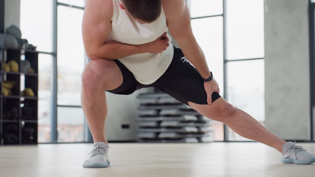 Gym junkie stretching knee in modern fitness studio, bending over in focused posture, dressed in white tank and black shorts, balancing on light wooden floor with windows in background