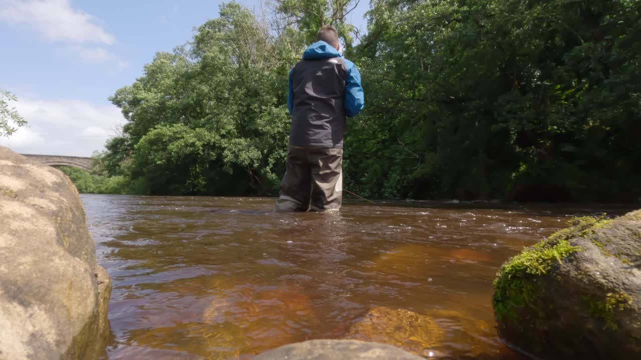 toma de ángulo bajo de un pescador vadeando y pescando con mosca en aguas marrones turbias