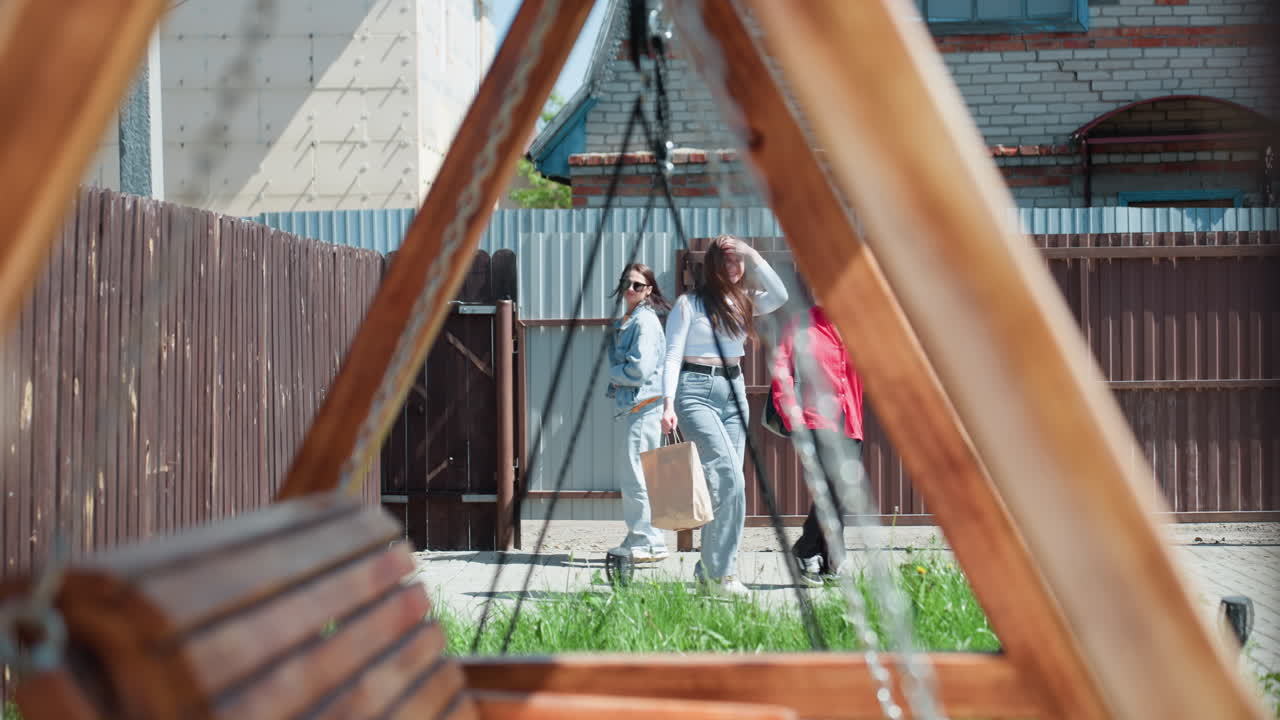 Wooden swing frame in sharp foreground with metal chains under sunlight, three girls visible in blurred background entering compound through open gate near tall fence