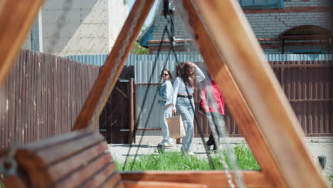 wooden swing frame in sharp foreground with metal chains under sunlight, three girls visible in blurred background entering compound through open gate near tall fence