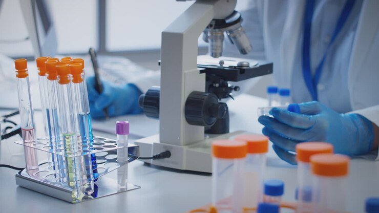Close Up Of Lab Worker Conducting Research Using Microscope Holding Test Tube Labelled Omega