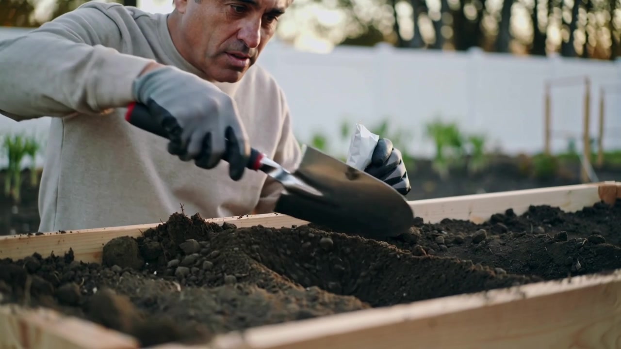 Hombre cavando con guantes grises, usando una paleta y sosteniendo un paquete, recogiendo tierra en una jardinera del patio para plantar.