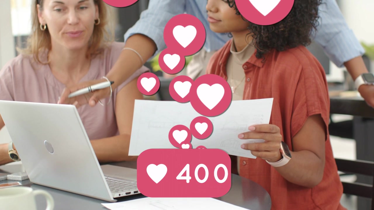 La líder se inclina señalando con un bolígrafo y corazones suben sobre el portátil mientras dos mujeres revisan el marketing.