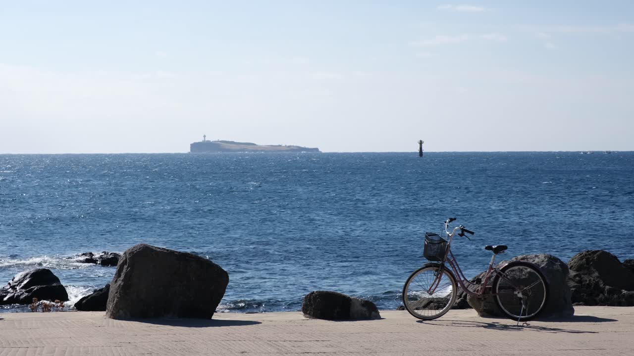 Pemandangan pantai yang tenang dari Pulau Marado dilihat dari Gapado, Jeju, dengan sepeda terparkir di hari yang cerah. Diambil dalam format lanskap, menampilkan laut yang tenang dan langit cerah