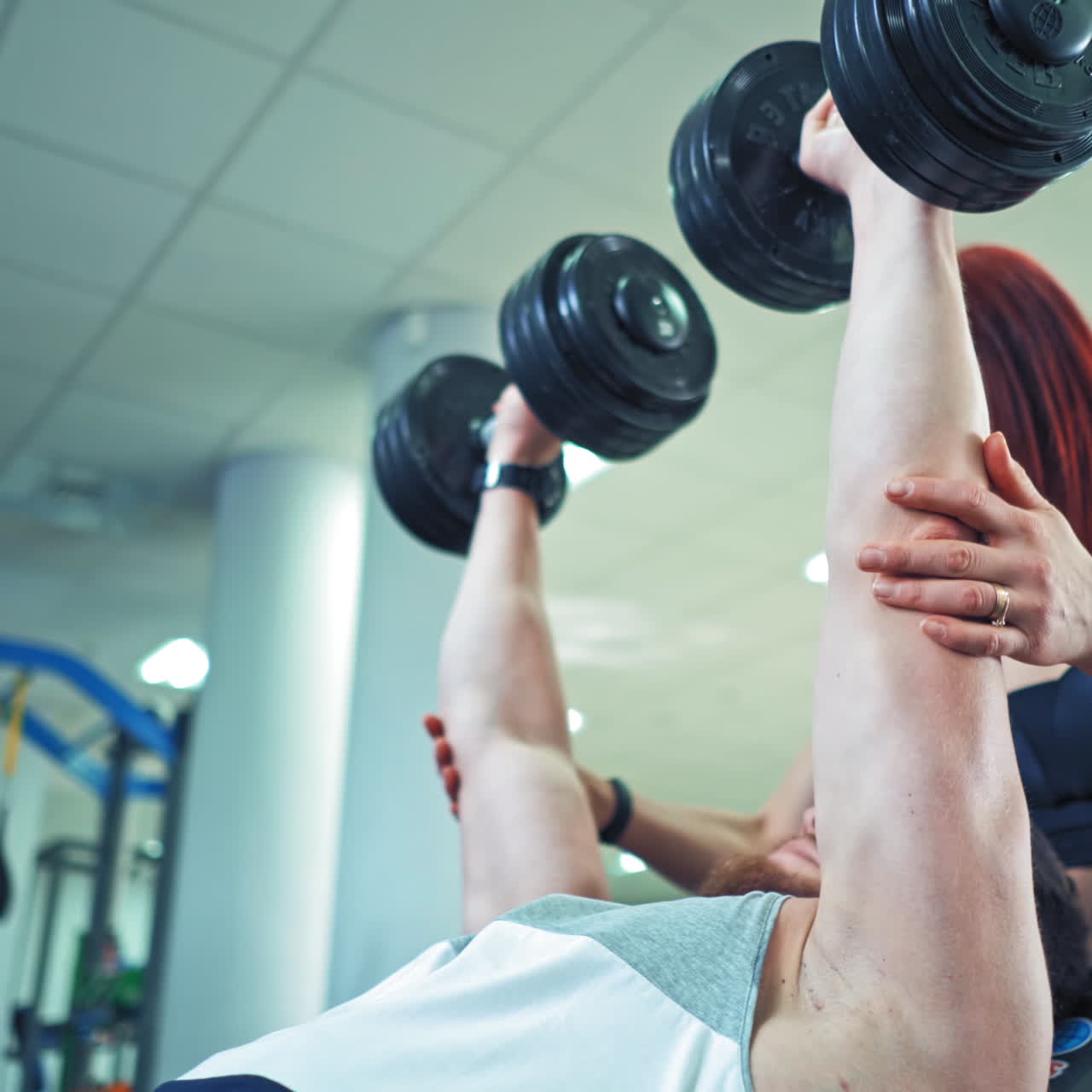 Smiling red-haired coach helps strong man to lift heavy dumbbells in sport club. Male athlete is lifting dumbbells while lying on desk. Personal trainer woman and a bodybuilder in gym.