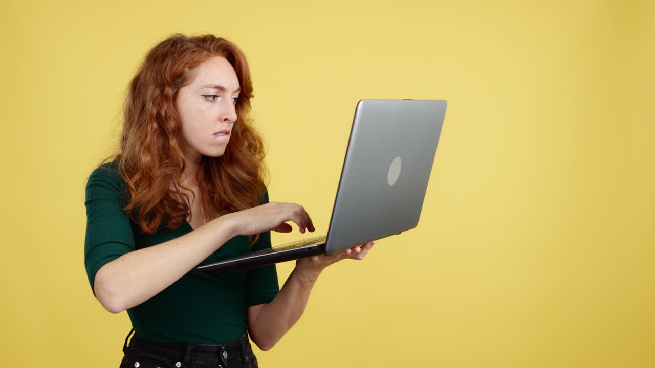 Red-haired woman using a laptop with a focused expression on a yellow background