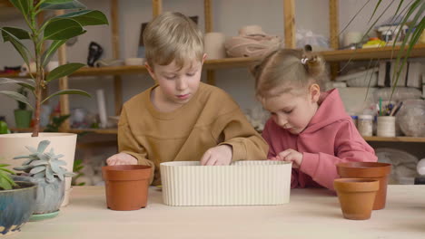 Kleines-Blondes-Mädchen-Und-Blondes-Kind-Bereiten-Den-Boden-In-Einem-Topf-Vor-Und-Sitzen-An-Einem-Tisch-Mit-Pflanzen-In-Einer-Handwerkswerkstatt-1