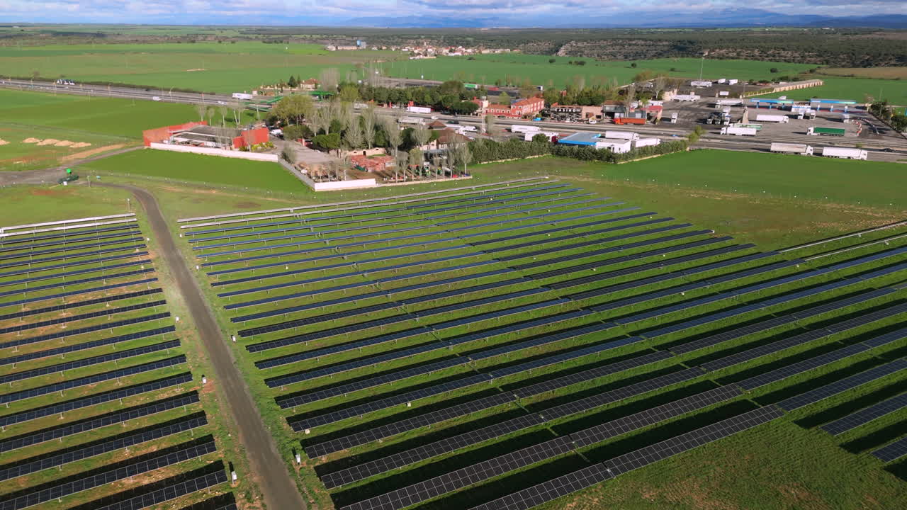 Aerial clip flying forward over rows of solar panels while gradually descending, showcasing a photovoltaic field and nearby industrial facilities under a clear sky