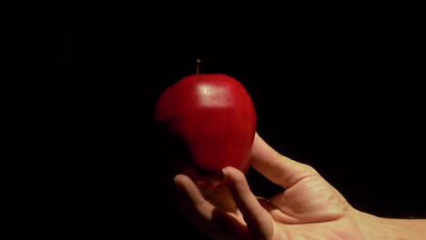 a white male hand comes into frame holding a bright red apple over a completely black backdrop