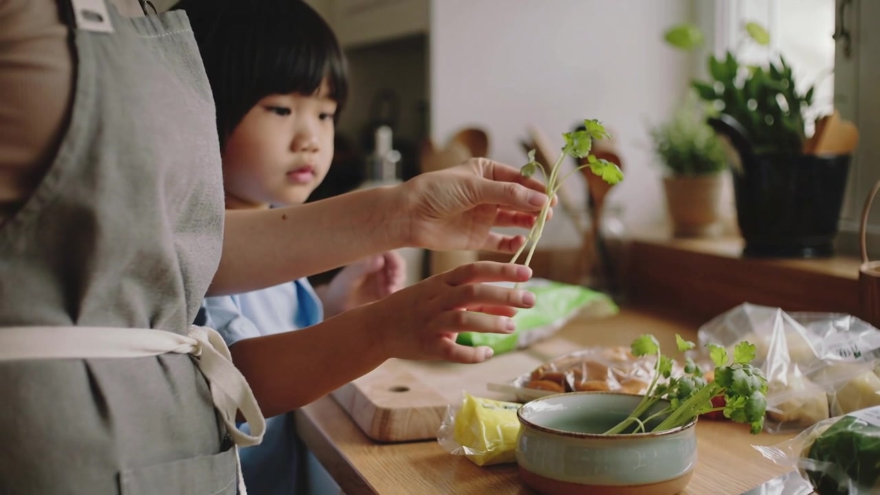 自宅のカウンターで、大人が子供に陶器のボウルからハーブの束を持たせるよう導き、食事の準備をする。