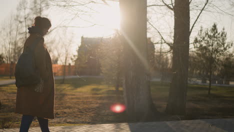 a girl wearing a brown coat, hoodie, and jeans walks alone in a park during sunset with her hands in her coat pockets, looking mournful,solitude and deep reflection, backdrop of bare trees