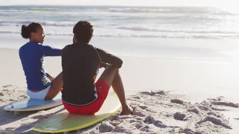 pareja afroamericana hablando y sentada de tablas de surf en la playa