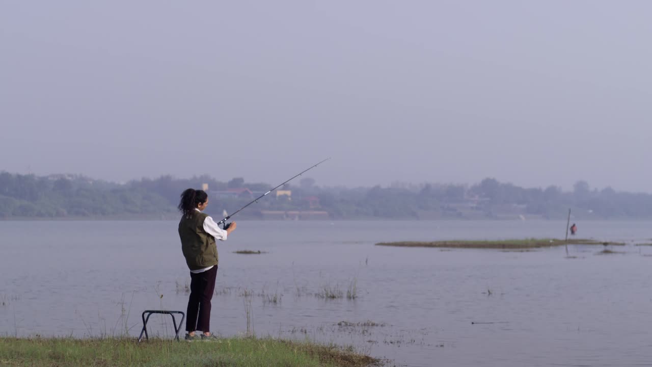 Seorang wanita Asia Selatan memancing di tepi danau yang tenang, berdiri di dekat kursinya dan melemparkan joran ke arah air terbuka sambil menikmati alam yang tenang di siang hari.