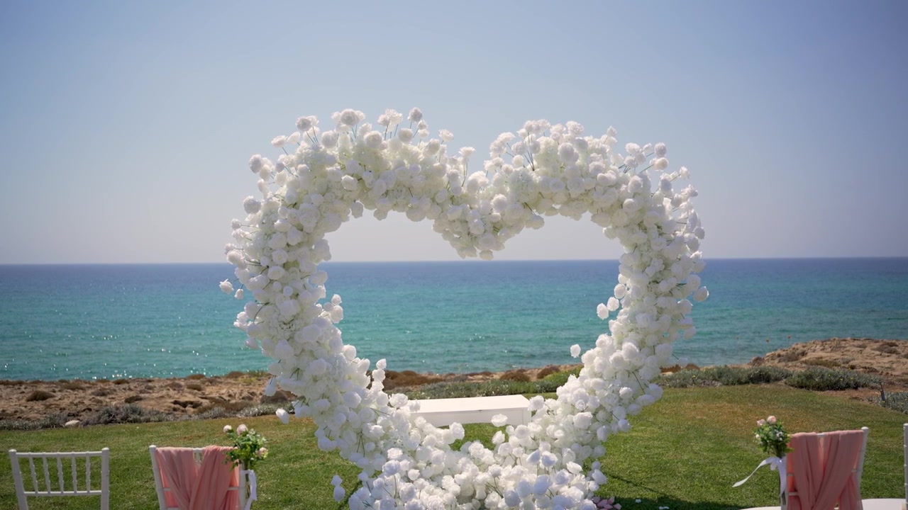 Et bryllup finder sted ved havet. Et stort hjerte lavet af blomster er opstillet på græsset. To hvide stole vender mod dekorationen under en klar himmel