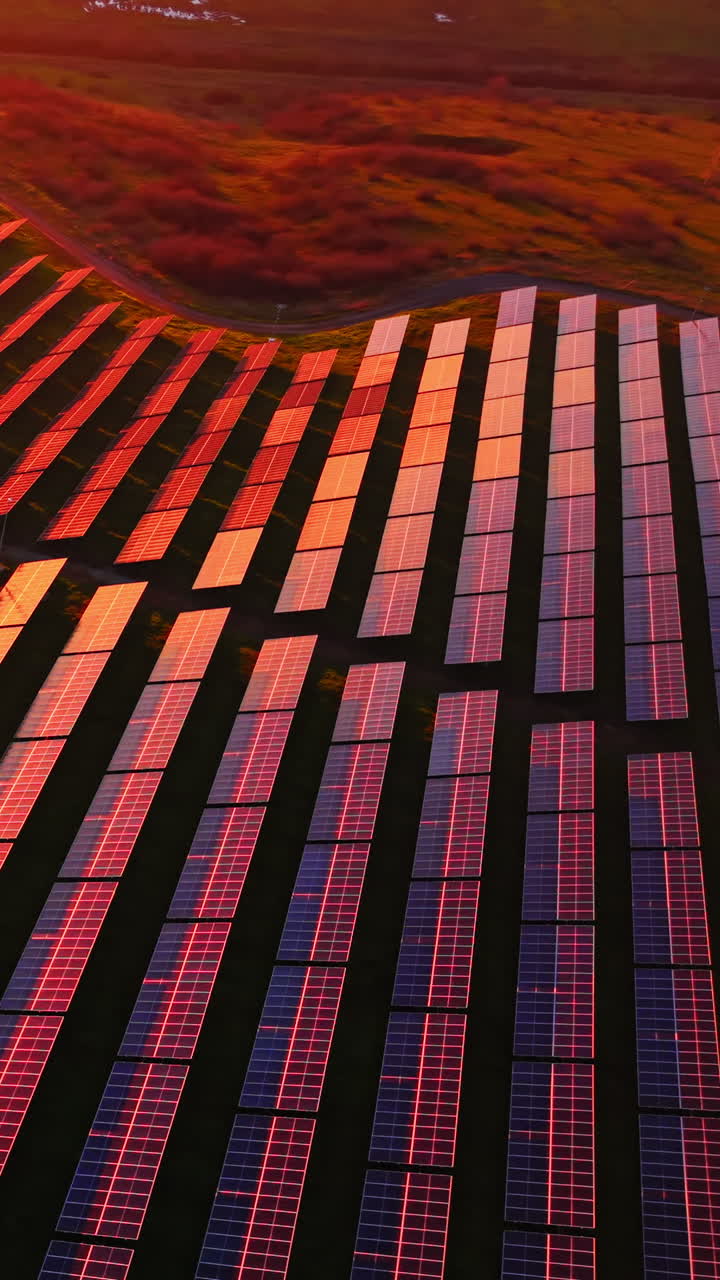 Vista dall'alto di impianti fotovoltaici su un paesaggio collinare sotto un'intensa illuminazione arancione e rossa del tramonto con ombre scure. Ripresa aerea di un parco solare. Video verticale