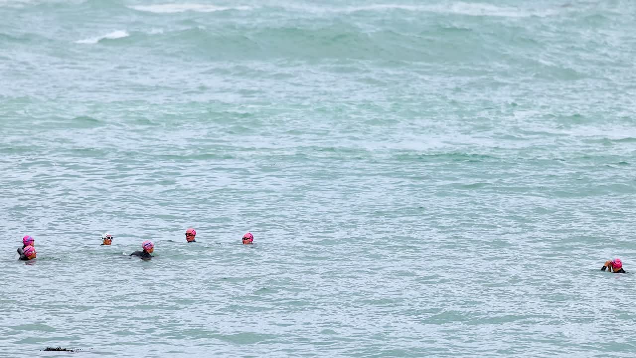 Swimmers in pink caps navigate ocean waves at Port Campbell, Australia. Overcast lighting and steady camera capture the serene seascape