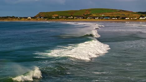 aerial tracking follows waves slowly crash and race to shoreline with village homes in background