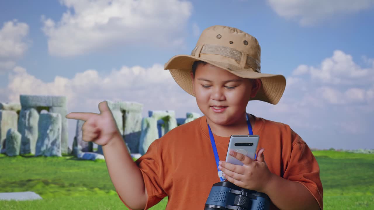 Asian Boy With A Hat And Binoculars Looking At Smartphone Then Smiling And Pointing To Side While Traveling In Stonehenge. Boy Researcher Examines Something, Travel Adventure, Close Up