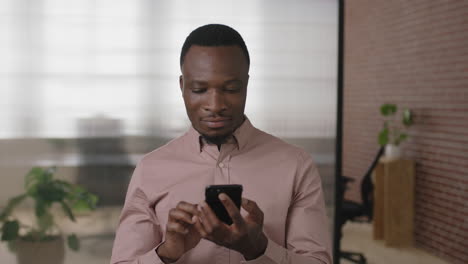 Porträt-Eines-Jungen-Afroamerikanischen-Geschäftsmannes,-Der-SMS-Schreibt-Und-Mit-Der-Smartphone-Netzwerk-App-In-Einem-Modernen-Büroarbeitsplatz-Surft