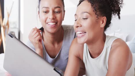 Happy-biracial-lesbian-couple-lying-on-bed-using-tablet,-in-slow-motion