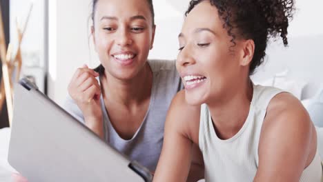 happy biracial lesbian couple lying on bed using tablet, in slow motion