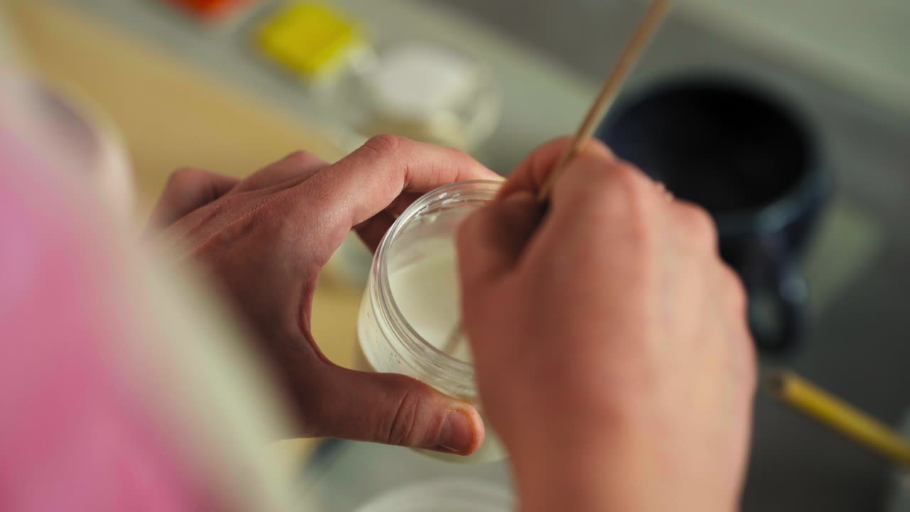 Artist's Hand Stirring Glaze In Small Jar Using Stick. Glazing Clay Cups At Workshop. high angle shot