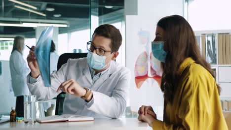 Médico-Caucásico-Con-Mascarilla-Médica-Sentado-En-El-Escritorio-Explicando-La-Enfermedad-Pulmonar-Por-Coronavirus-A-Una-Paciente-En-Consulta-Médica