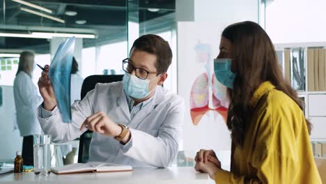médico caucásico con mascarilla médica sentado en el escritorio explicando la enfermedad pulmonar por coronavirus a una paciente en consulta médica