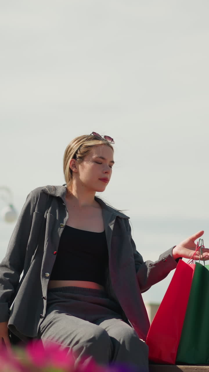 여자는 편안하게 그녀의 쇼핑 가방을 벤치에 떨어뜨리고 편안한 표정으로 앉아서 멀리 쳐다보고, 빛의 기둥과 흐릿한 초록색이 배경에 보입니다.