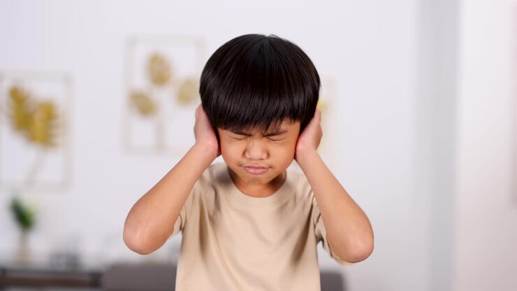 Young Asian boy covers his ears and displays discomfort in a softly lit, modern room with neutral decor, captured in a steady medium shot