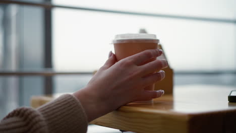 primer plano de la mano de la dama tocando la taza de café en la mesa de madera con fondo borroso suave, vista parcial del teléfono visible, entorno de café moderno con luz natural, momento de relajación con bebida caliente