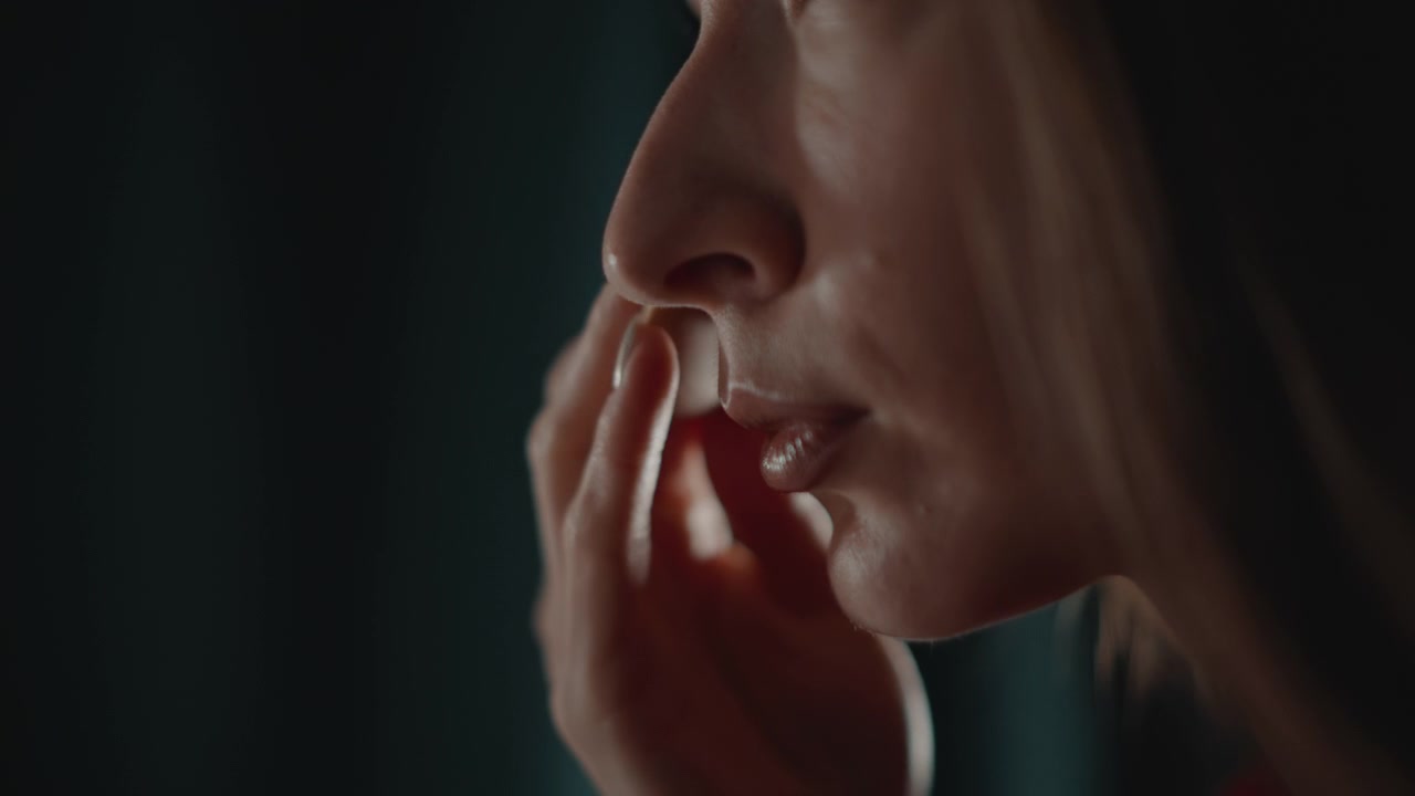 Vista di profilo di una donna che preme una spugnetta per il trucco lungo la mascella mentre le mani tengono un oggetto rettangolare sottile nelle vicinanze, con un leggero movimento della bocca, dita posate, luce soffusa e cura della bellezza concentrata.