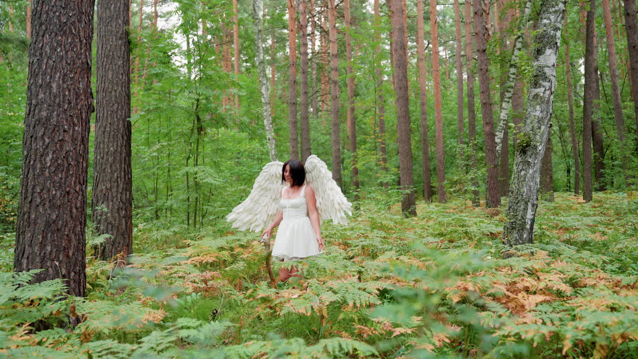Actress dressed like angel walking through peaceful forest with wooden staff, surrounded by tall trees and lush ferns, wearing white dress with large wings, exploring tranquil green woodland scenery