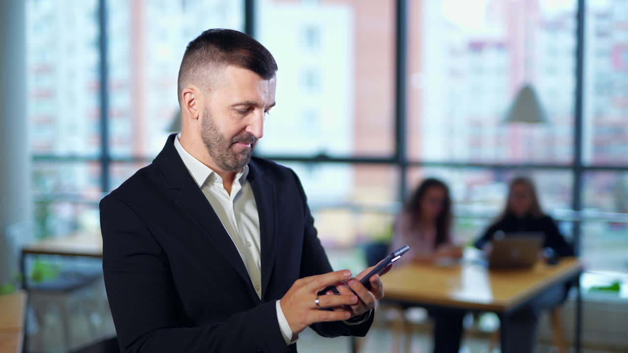 Businessman standing in office and holding phone. Entrepreneur messaging to somebody. Portrait close up. Females in blur at backdrop.