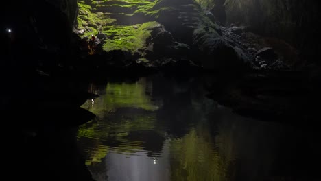 Panorámica-Hacia-Arriba-Desde-El-Reflejo-En-El-Agua-Hasta-La-Dolina-Con-Rayo-De-Sol-Dentro-De-La-Cueva-De-Son-Doong,-Siluetas-De-Personas-Moviéndose