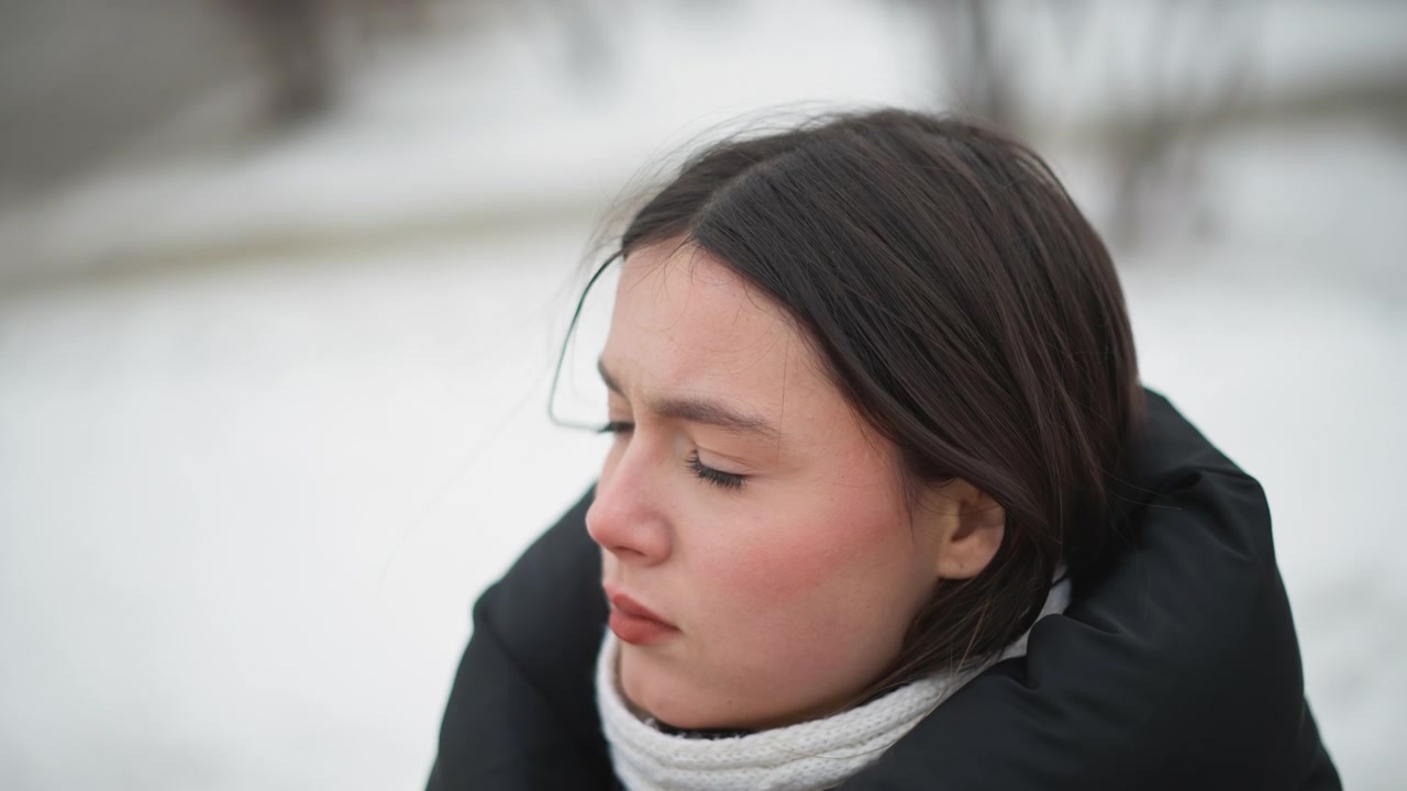 Close up of woman wearing black coat and white scarf in snowy outdoor setting. Subject remains still with minimal movement maintaining consistent expression. Background shows blurred snow