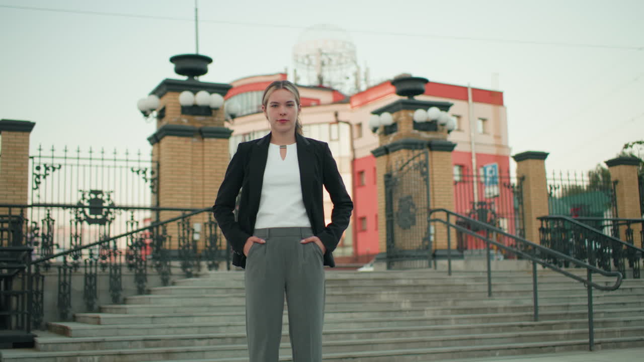 Young woman confidently poses with hands on waist while standing on steps in urban area with modern buildings and metal railings behind, expressing assertiveness in professional attire