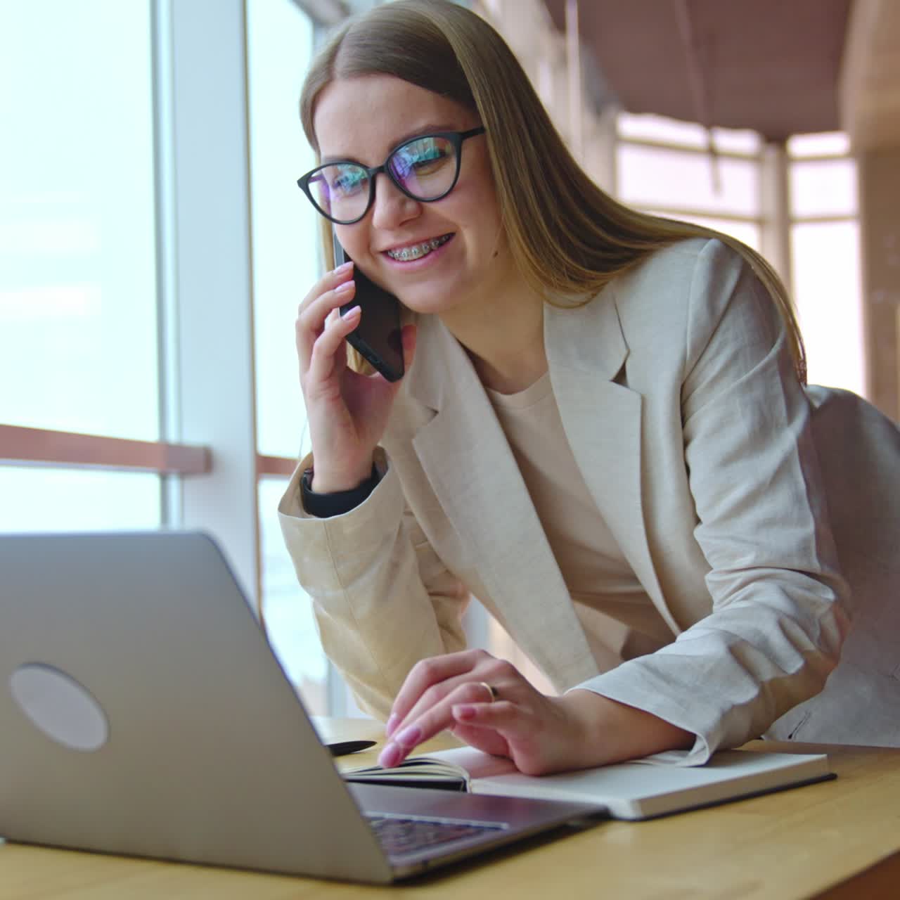 Cheerful positive long-haired lady in glasses talks on the phone standing bent over the desk. Woman presses keys on her laptop during conversation
