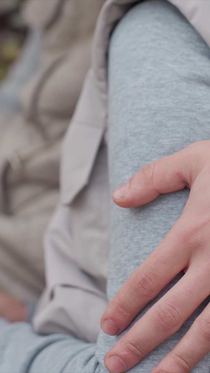 Le dita si incurvano leggermente mentre poggiano su un materiale grigio. La texture dei pantaloni grigi è uniforme sotto un leggero peso. Una telecamera stabile cattura un primo piano, mantenendo la messa a fuoco senza movimento.