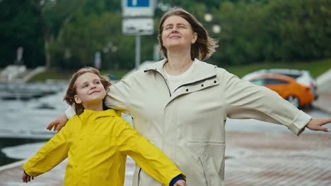 Mujer-Rubia-Feliz-Junto-Con-Su-Hija-Adolescente,-Una-Niña-Con-Una-Chaqueta-Amarilla,-Se-Paran-Contra-El-Viento-Y-Disfrutan-Del-Viento-Mientras-Caminan-En-El-Parque-Después-De-La-Lluvia