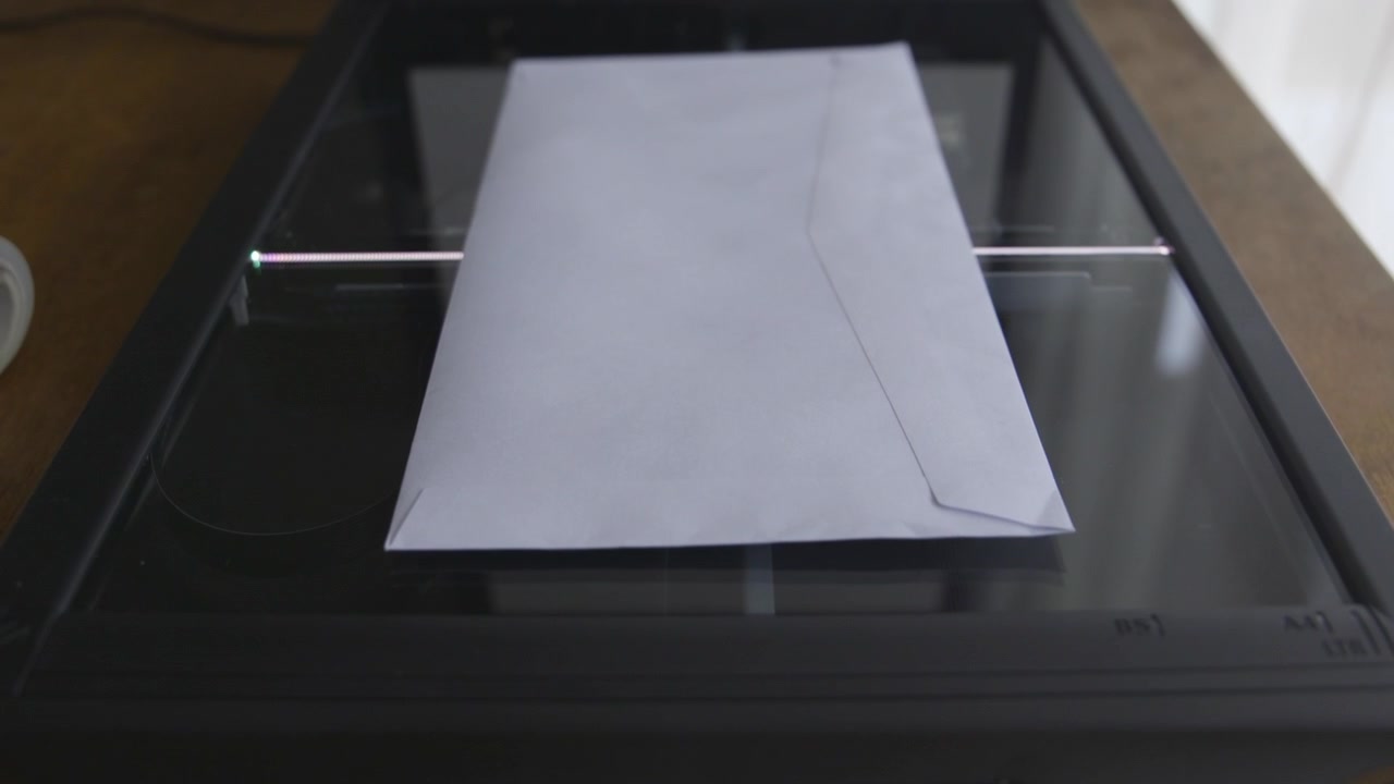 Bílá obálka je položena na skleněný povrch plochého skeneru, připravená ke skenování. Ideální pro témata kancelářské technologie nebo digitalizace dokumentů.