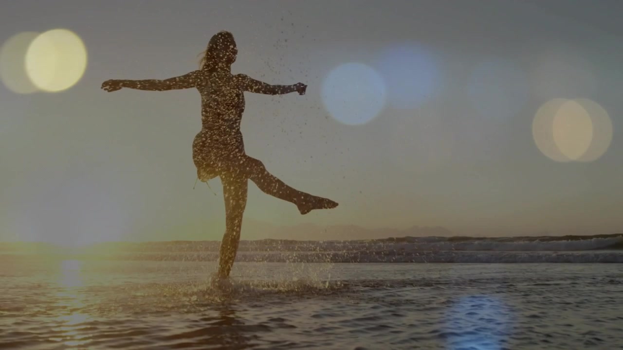 Woman walking sunset surf, lifting leg, kicking water, creating sparkle, lens flare for travel