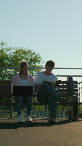 estudiantes universitarios estudiando al aire libre con una computadora portátil en el regazo de la chica mientras el chico toma notas, ella revisa su trabajo, rodeados de vegetación, postes de luz y bancos de madera bajo un ambiente sereno y soleado