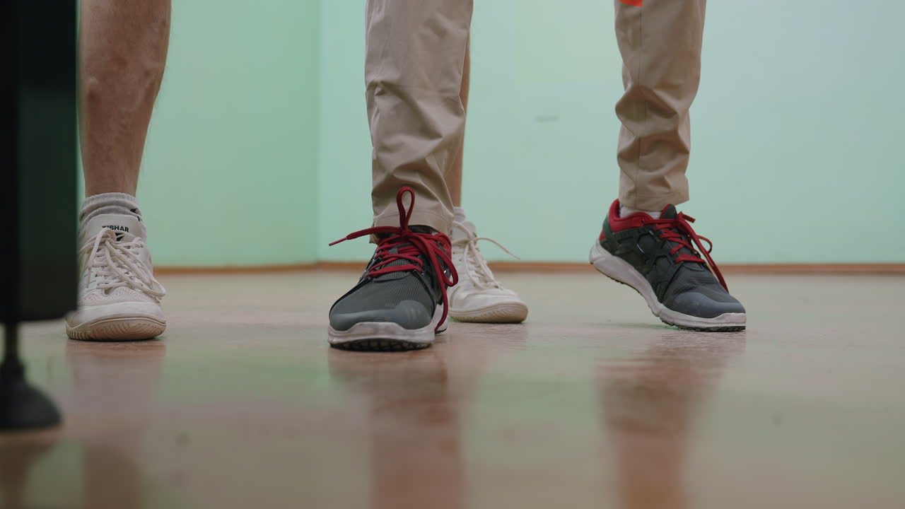 Leg view of people moving as if dancing on polished indoor floor while tennis player practices behind them in sports hall, blending playful movement with athletic training atmosphere