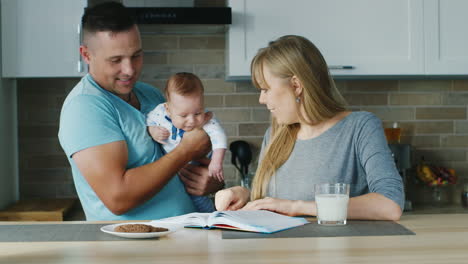 young family in the kitchen mom reading a book with the baby daddy is near