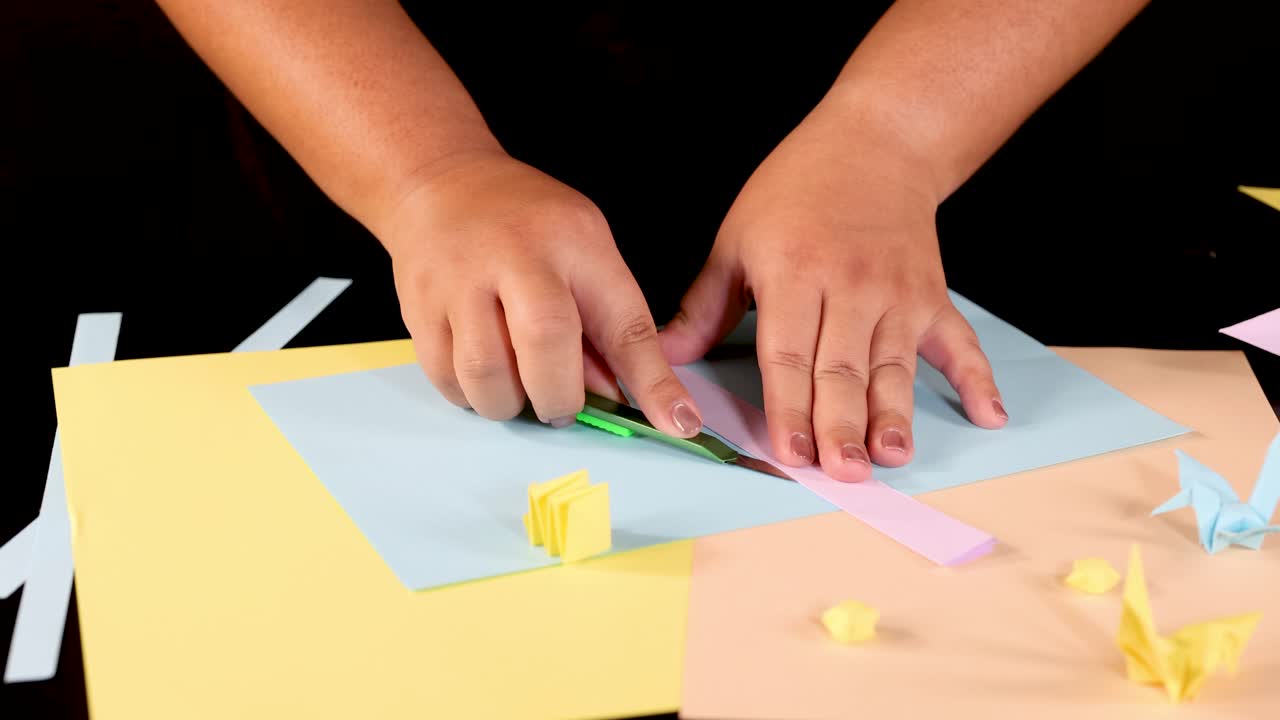 Person folds pastel paper strips on table, creating origami shapes under bright, even lighting