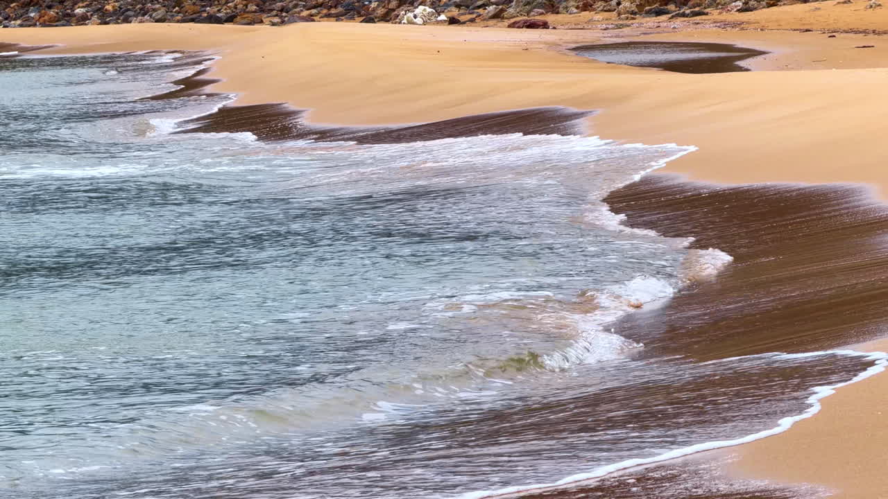 Onde che si infrangono delicatamente sulla riva sabbiosa in una spiaggia tranquilla durante il giorno. Le onde si muovono sulla sabbia di una spiaggia durante il giorno.