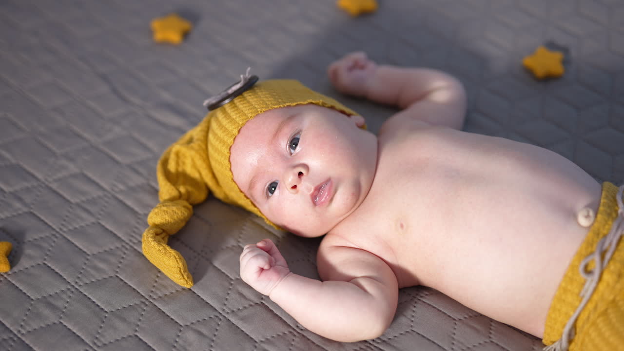Infant boy with naked belly lying still on the bed. Baby in a yellow cap and pants looks intensely into the camera. Close up.