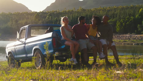 friends with backpacks sitting on tailgate of pick up truck on road trip by lake in countryside