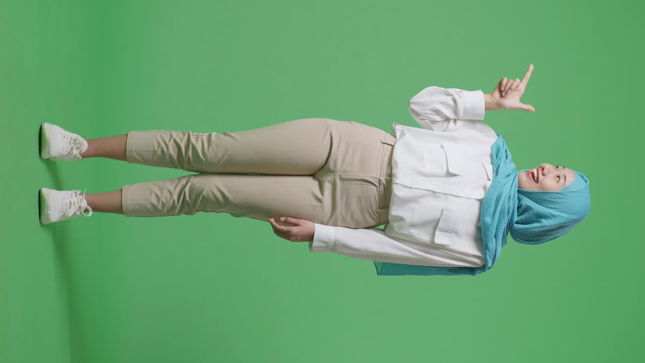 녹색 스크린 배경 스튜디오에 서 있는 동안 웃고 을 가리키는 아시아 무슬림 여성의 몸이 가득 차 있습니다.